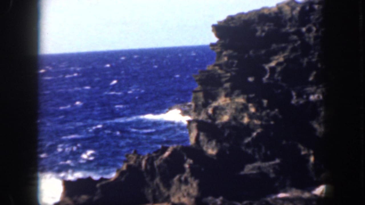 1959 Oahu shoreline frame with rugged dark rock, deep blue ocean, white water, and a bright sky beyond the cliff edge.