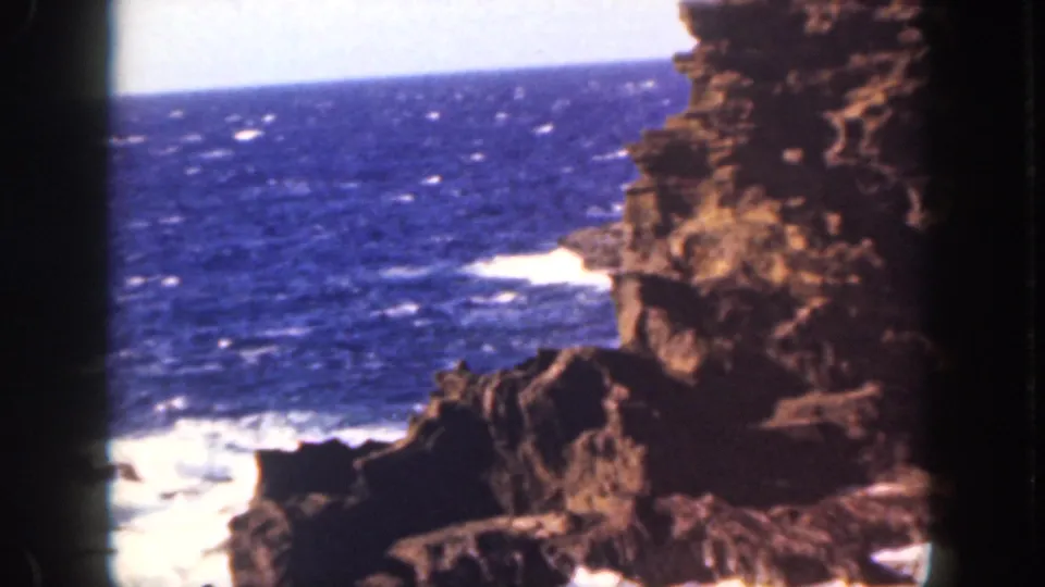 1959 Oahu coastal frame showing dark rocks, foamy surf, and blue ocean with the warm color cast of scanned 8mm film.