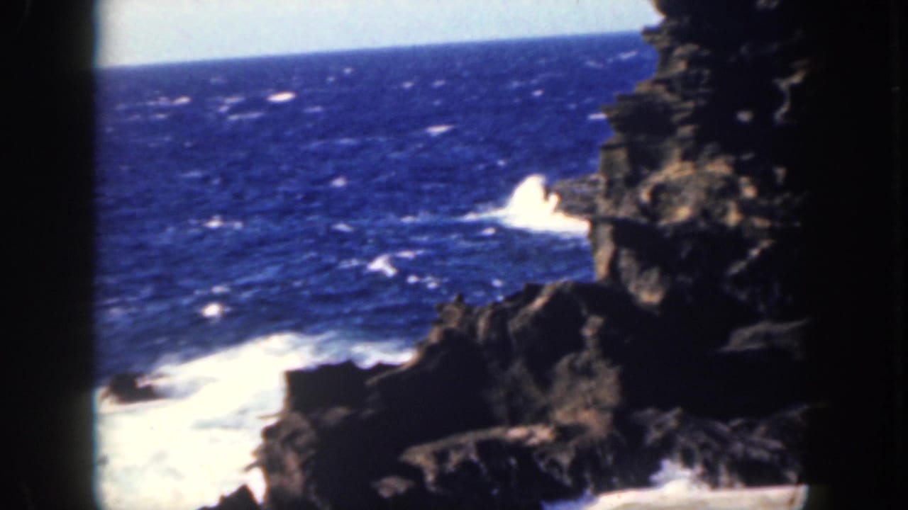 Blue ocean waves crash against dark volcanic rocks seen through a shadowed rock opening.