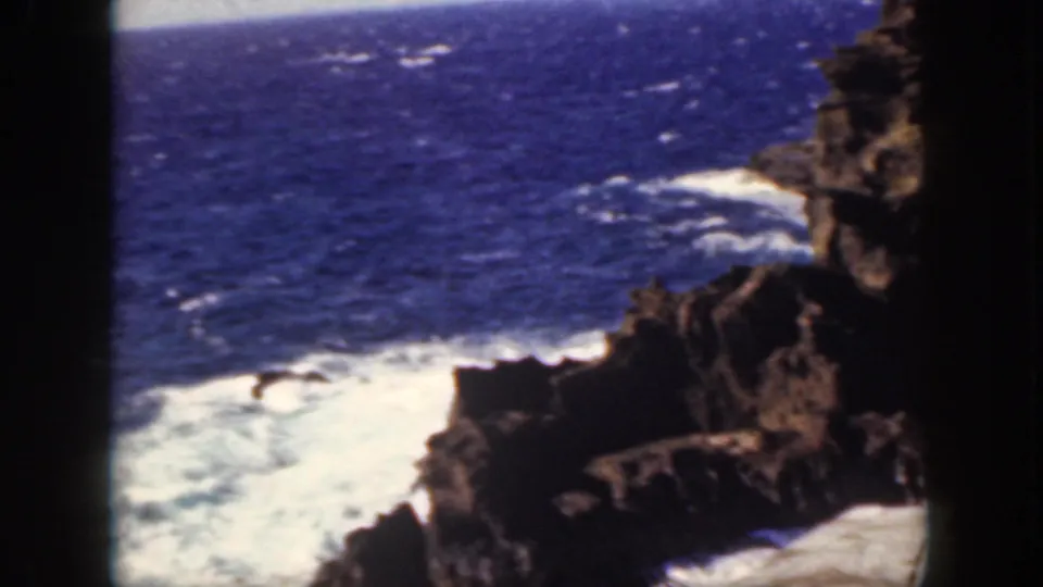 1959 Oahu shoreline view with foaming waves moving around dark volcanic rocks under a blue sea horizon.