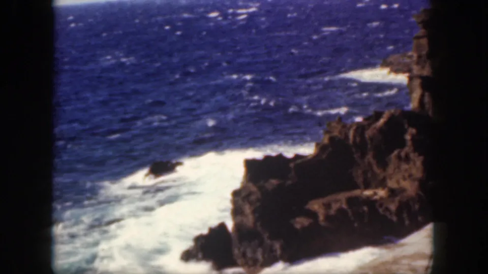 1959 Oahu 8mm frame of surf rolling around jagged shoreline rocks, with blue water beyond and a shadowed foreground edge.