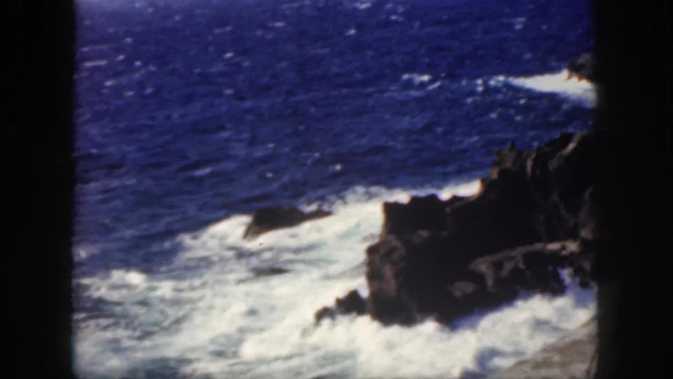 1959 Oahu coastal frame showing white surf breaking against dark volcanic rock, with open blue ocean in the background.