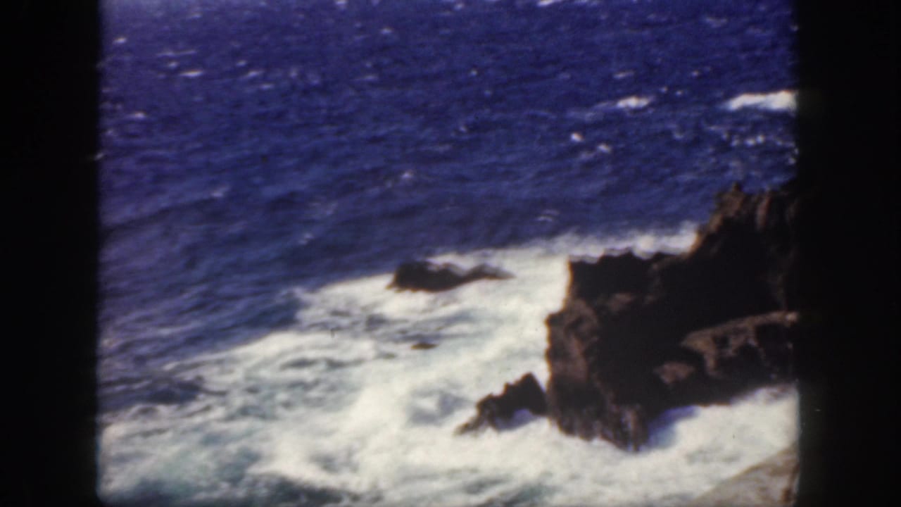 1959 Oahu 8mm frame showing white Pacific surf breaking against dark volcanic rocks under deep blue water.