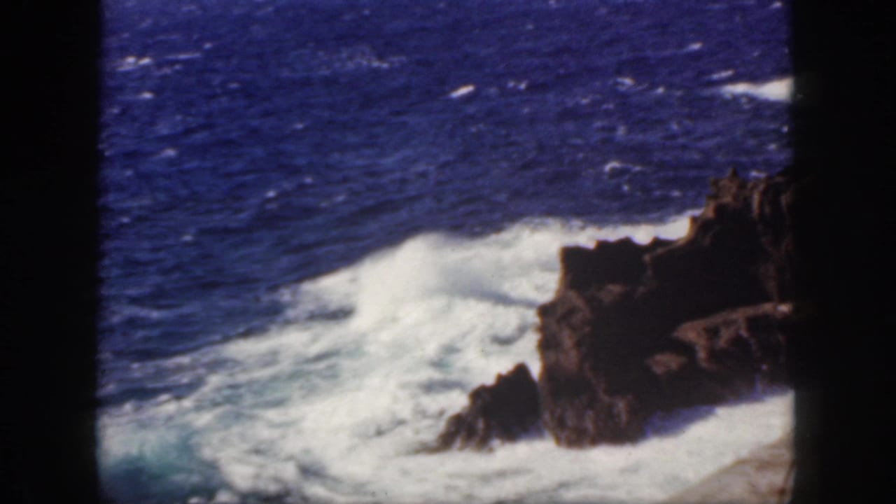 1959 Oahu coastal frame with a foaming wave wrapping around dark rocks in a saturated blue ocean.