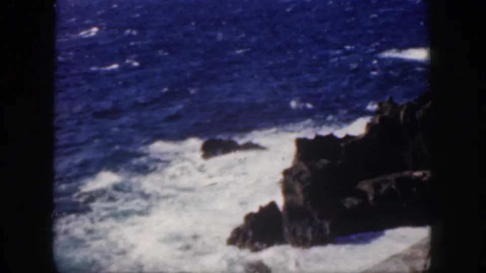 1959 Oahu 8mm frame showing white foam and churning surf against a rocky volcanic shoreline.