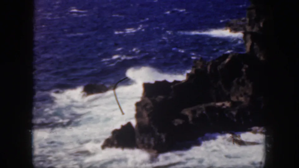 1959 Oahu shoreline frame with surf washing across dark rocks and open blue ocean in the background.