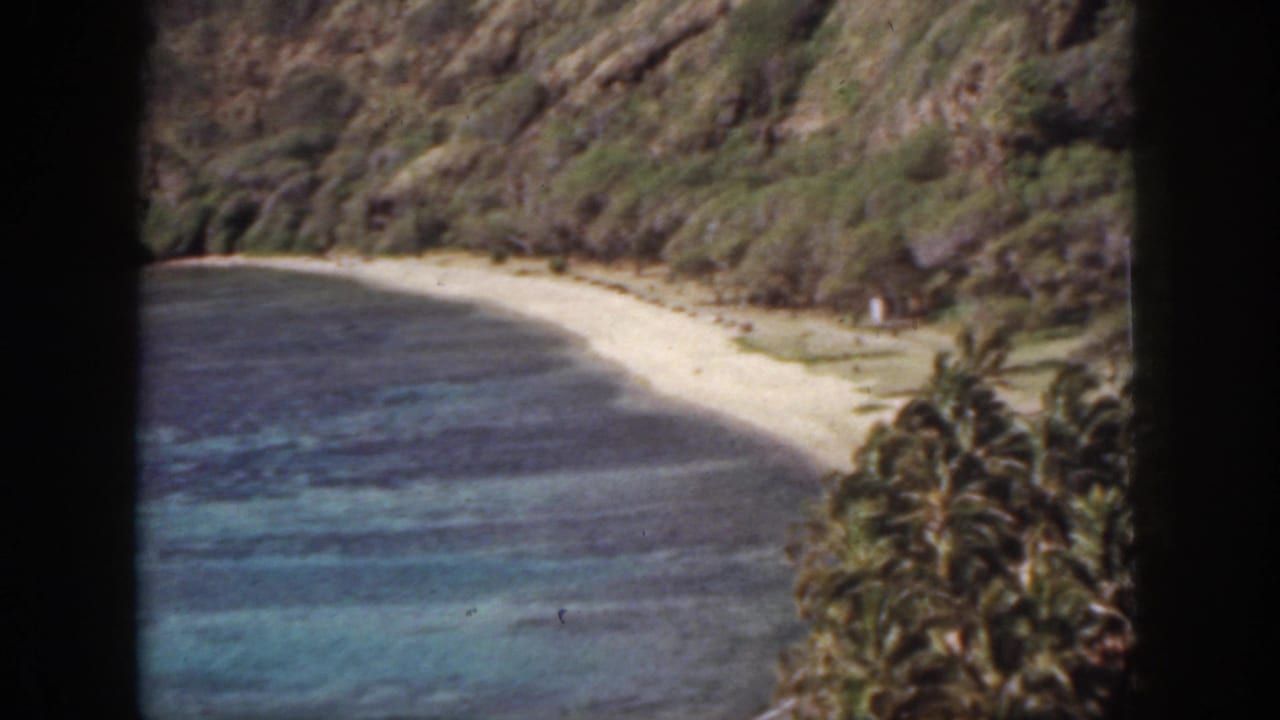 Curving sandy beach beside dark blue water, framed by palms and steep hills.