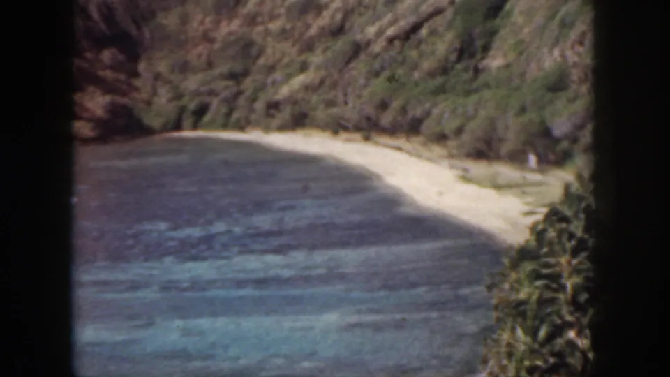 An elevated 1959 Oahu home-movie frame shows pale sand, blue-green reef water, rocky slopes, and palm tops near the beach.