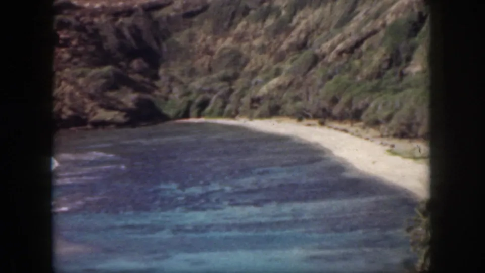 1959 Oahu 8mm view of a sheltered cove, showing reef-colored water, a narrow sandy beach, rugged slopes, and palms at right.