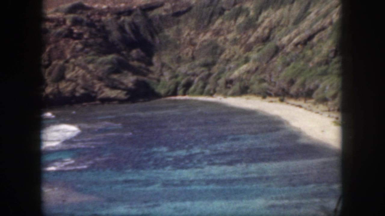 1959 Oahu home-movie frame showing a secluded cove with blue-green water, pale sand, and steep rocky cliffs.