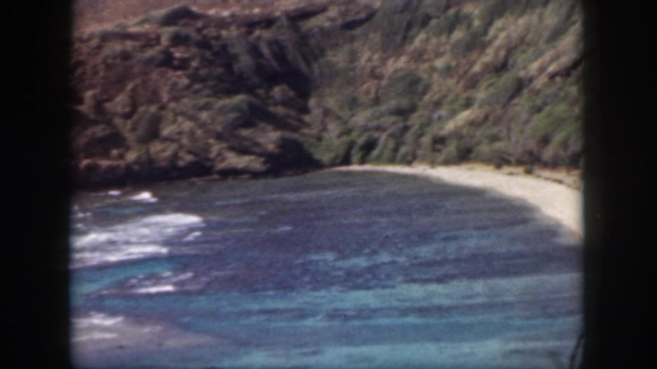 1959 Oahu shoreline view with a curved pale beach, breaking waves, and vegetation on rocky cliff faces.