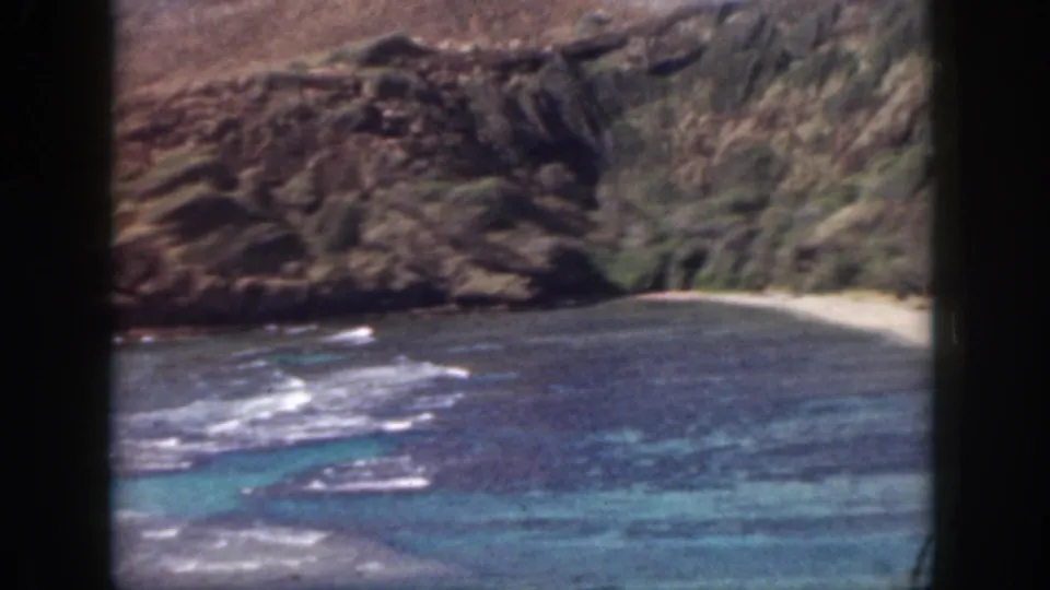 1959 Oahu coastal frame showing white surf over blue-green water, a small beach, and rugged cliff walls.