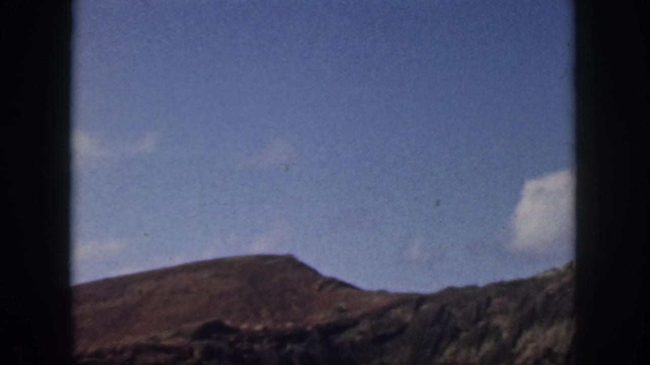 1959 Oahu home-movie frame showing blue sky, scattered clouds, and a rugged hillside seen through dark window-like borders.