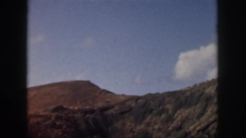 1959 Oahu home-movie view of rugged brown terrain and scrub-covered slope beneath a blue sky and bright cloud.