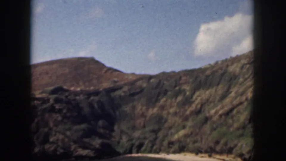 1959 Oahu landscape frame showing rocky volcanic-looking slopes, scrub vegetation, blue sky, and a white cloud within dark film borders.