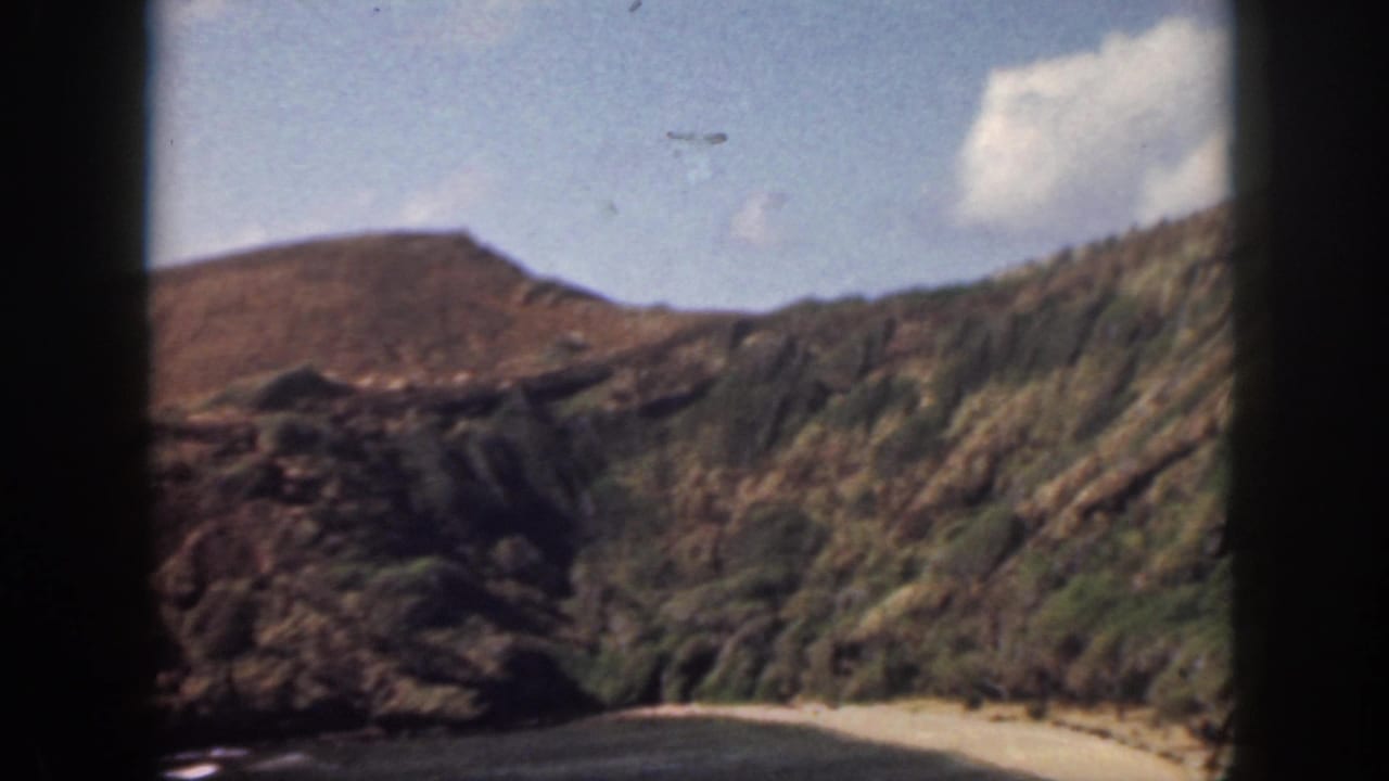 1959 Oahu home-movie frame showing rugged coastal hills, rocky cliffs, beach, ocean, and dark car-window framing.