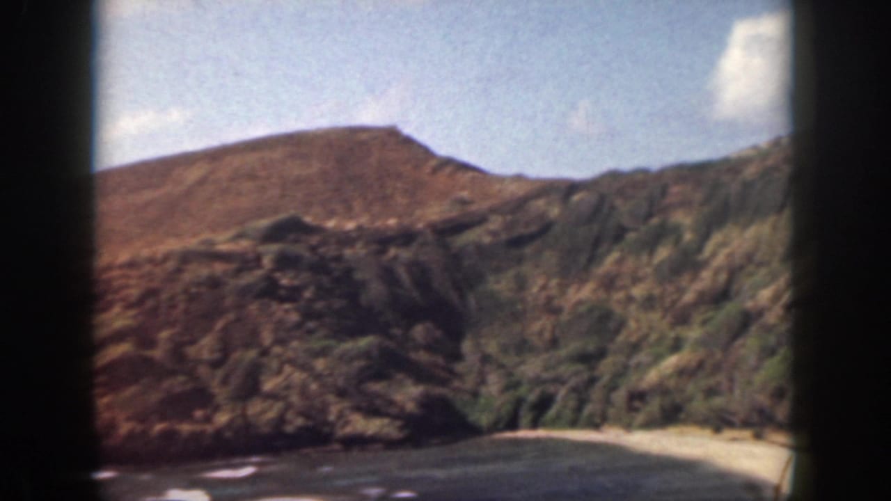 1959 Oahu coastal view with sunlit slopes, rocky cliffs, sandy shoreline, ocean water, and black window borders.