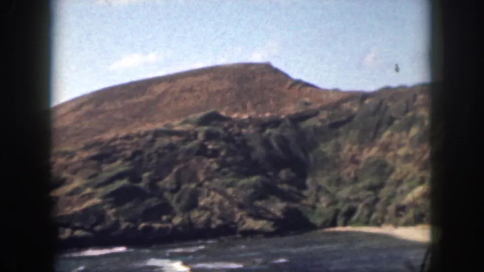 1959 Oahu frame showing a broad brown ridge, darker coastal rocks, green vegetation, ocean, and vehicle-window framing.