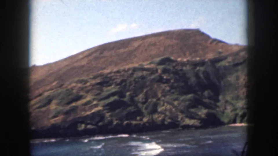 1959 Oahu home-movie still with a sunlit hill, rocky coastal outcrop, blue water, small surf, and dark frame edge.