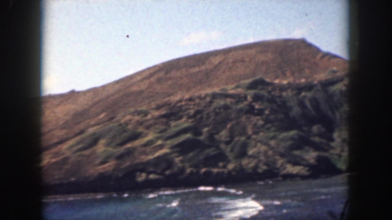 1959 Oahu home-movie frame showing a sun-baked hillside descending to dark coastal rock and breaking waves.