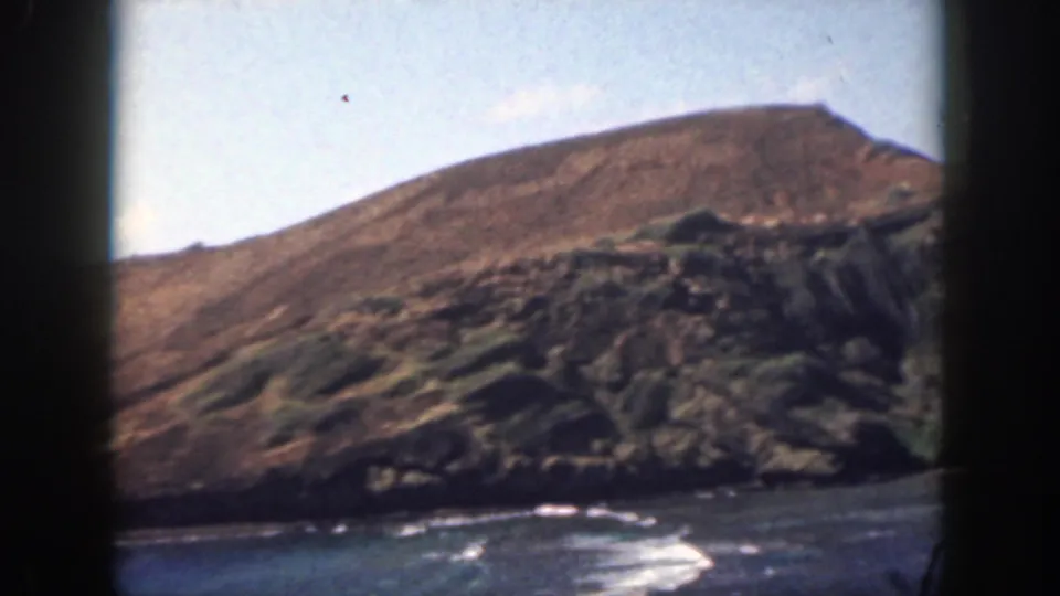1959 Oahu home-movie frame showing a sun-baked hillside descending to dark coastal rock and breaking waves.