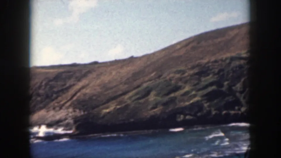 1959 Oahu home-movie still of a sloping coastal hillside, rocky edge, and blue water with small white breakers.