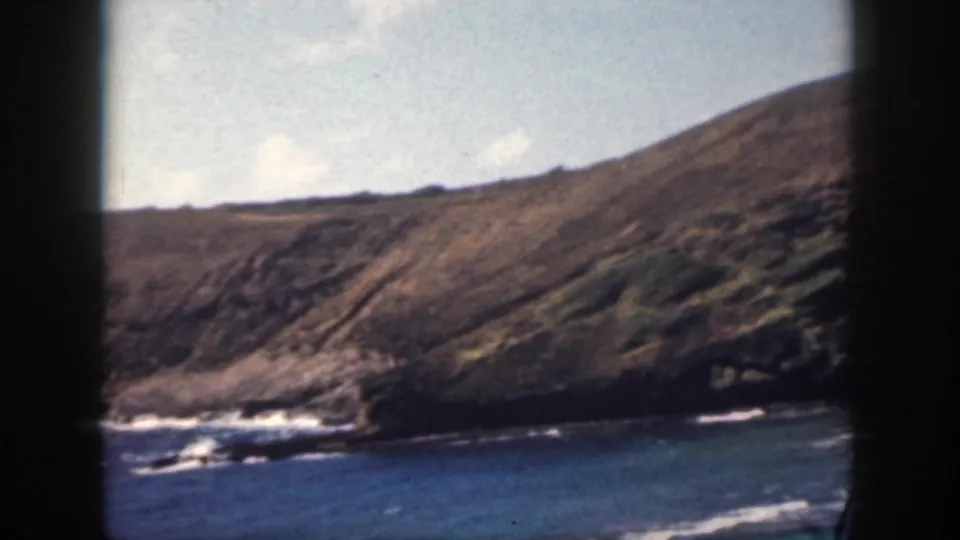 1959 Oahu frame showing rugged hillside terrain, dark shoreline rock, and blue ocean water with surf marks.