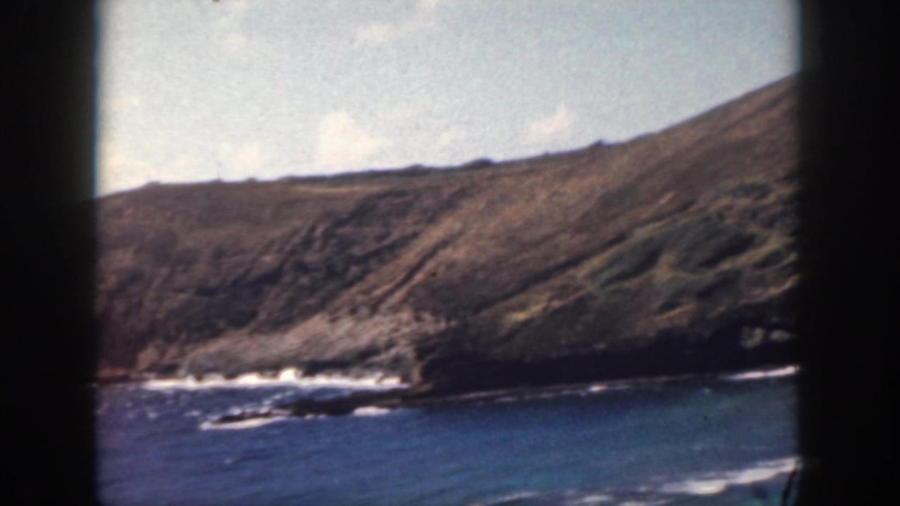 1959 8mm home-movie frame of an Oahu rocky shoreline, with blue ocean water, breaking surf, and a brown-green cliff under pale clouds.