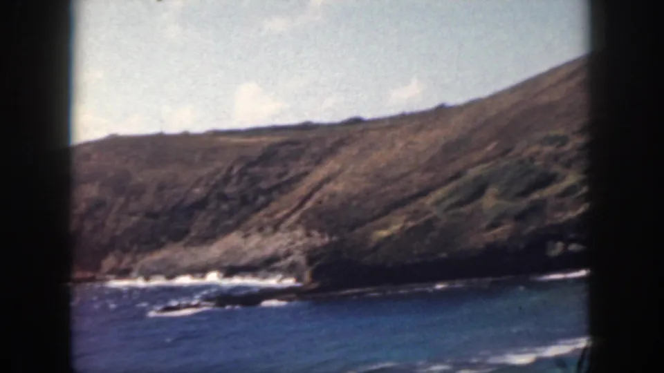 1959 8mm home-movie frame of an Oahu rocky shoreline, with blue ocean water, breaking surf, and a brown-green cliff under pale clouds.
