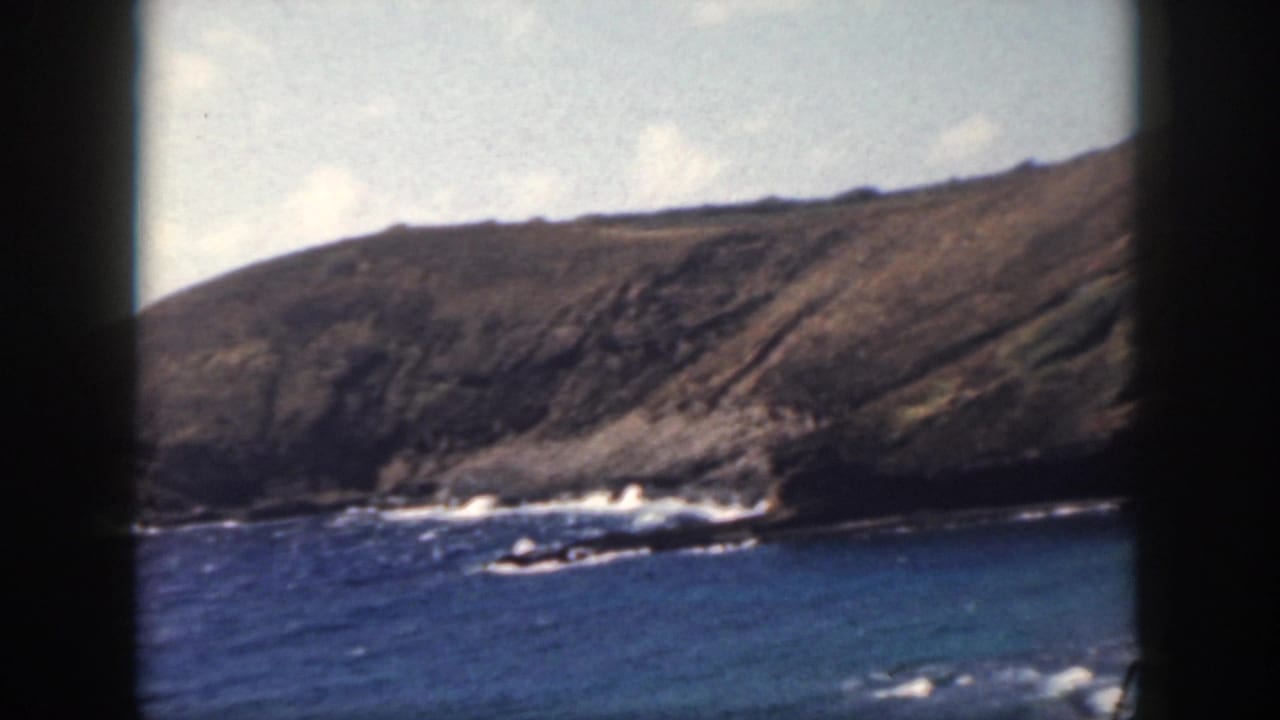Blue ocean and white waves along a rocky shoreline beneath a sloping coastal cliff.