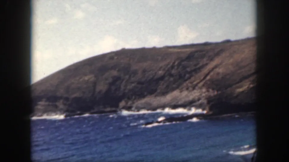 1959 home-movie view from Oahu with choppy blue water in the foreground, surf along rocks, and a sunlit coastal hillside beyond.