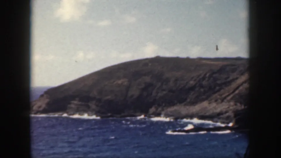 1959 8mm frame of an Oahu shoreline, showing a rounded coastal headland, dark rocks, blue water, and scattered surf foam.