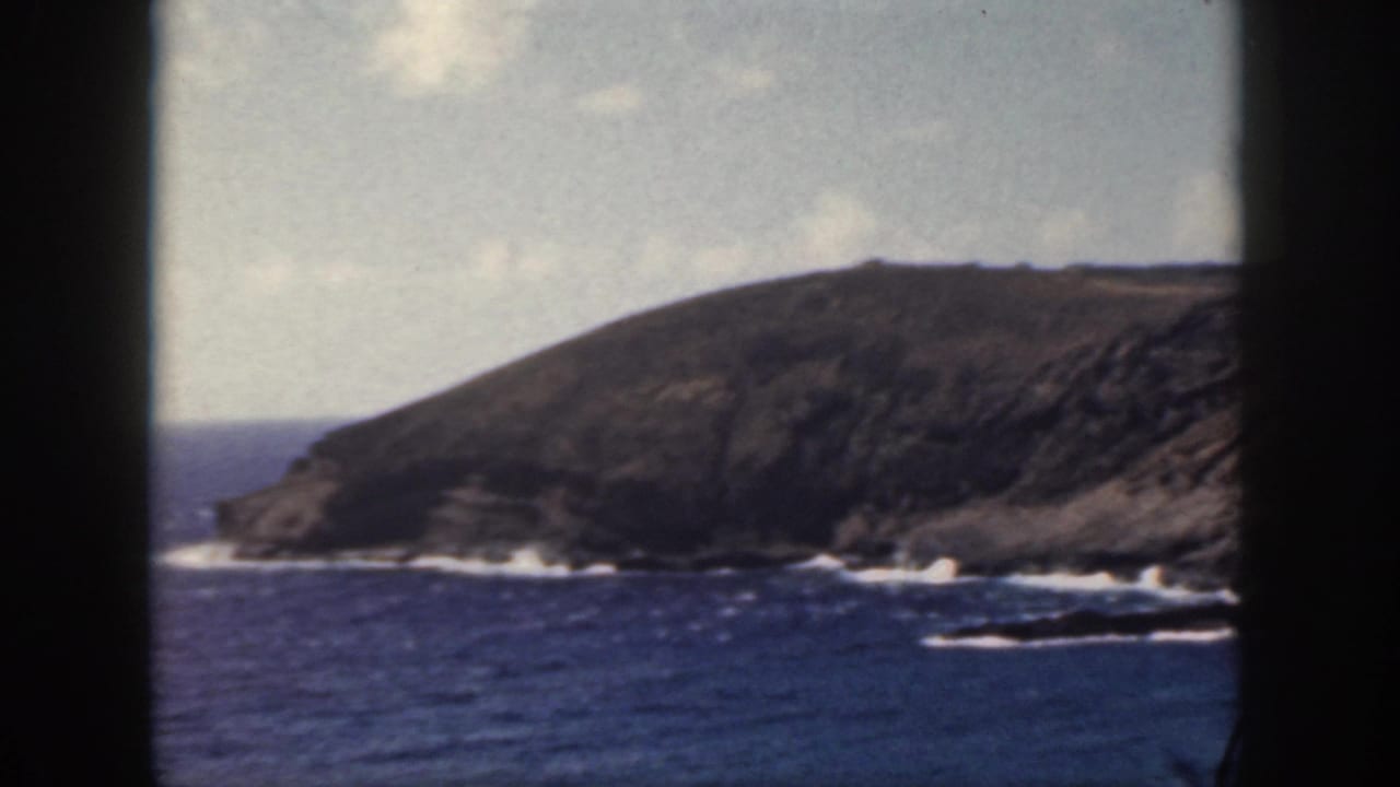 1959 Oahu 8mm frame showing a dark rocky headland, blue ocean, white breaking waves, and a pale cloudy sky.