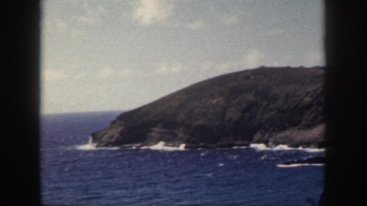 1959 Oahu coastal view with a rugged headland, blue water, white surf lines, and visible 8mm frame borders.