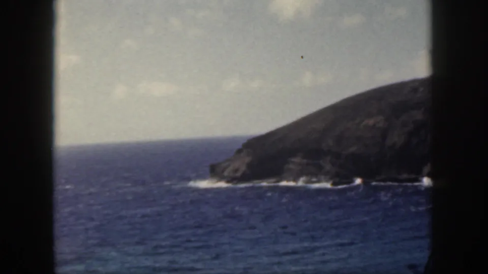 1959 Oahu coastal frame with a rocky headland, blue sea, white surf, and dark viewfinder-like borders.