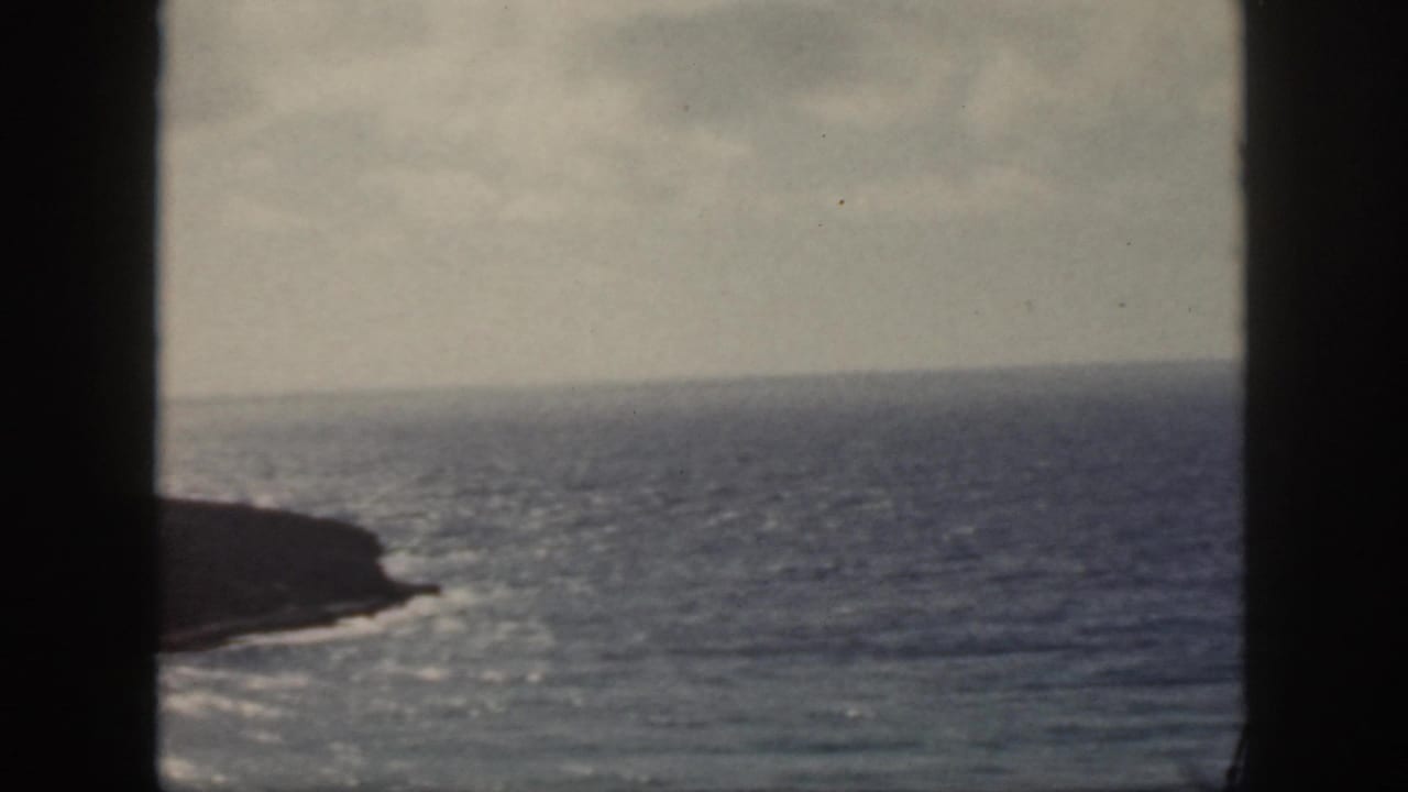1959 Oahu home-movie frame showing blue ocean, a dark rocky coast, pale sky, and vertical foreground framing.