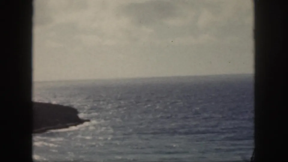 1959 Oahu home-movie frame showing blue ocean, a dark rocky coast, pale sky, and vertical foreground framing.