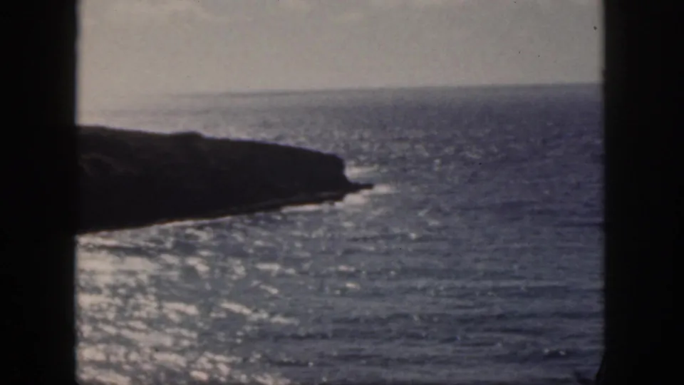 1959 Oahu 8mm frame with dark rocky shore, calm blue water, distant horizon, and foreground window-like framing.
