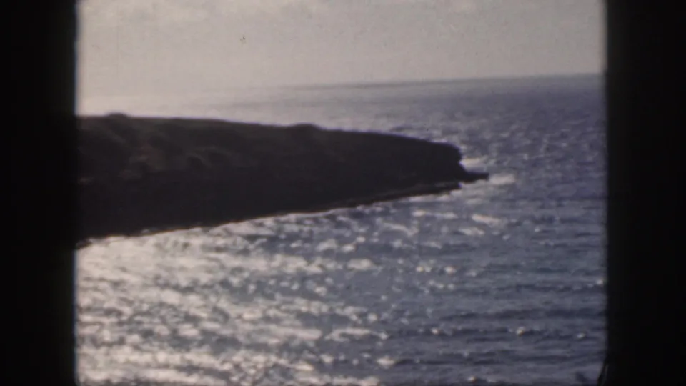 1959 Oahu coastal frame showing a dark rocky point against sunlit ocean, with pale sky and 8mm softness.