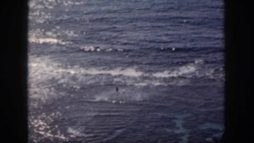1959 Oahu 8mm frame with sunlit ocean, low waves, and a dark gate border dividing the view.