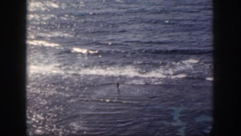 1959 Oahu 8mm frame showing a distant person wading in shallow surf near a rocky shoreline.