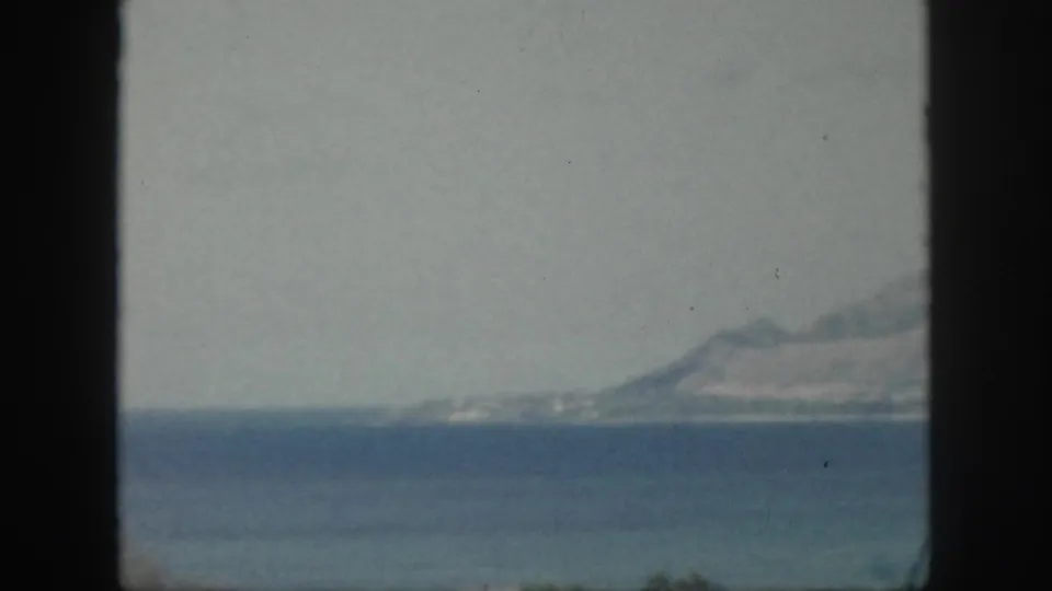 1959 Oahu home-movie view across blue water toward a hazy coastline and mountains, partly blocked by a dark vertical window frame.