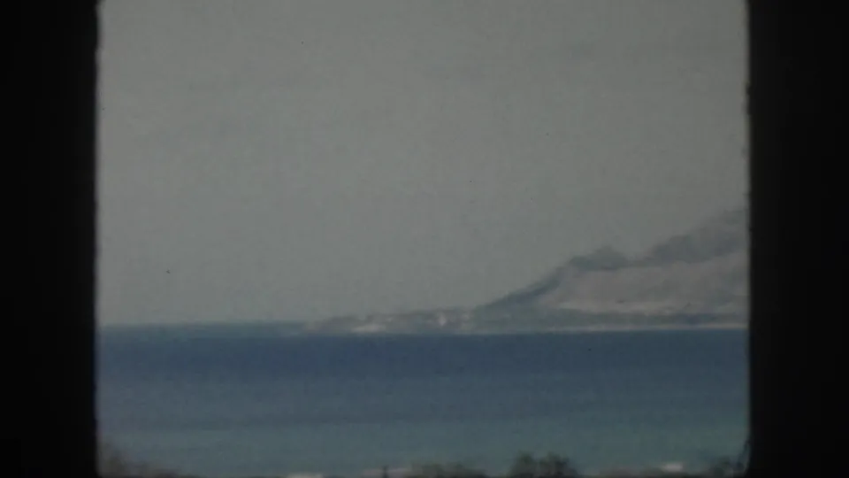 1959 Oahu 8mm frame with a hazy mountain shoreline seen beyond layered blue water and a pale sky.