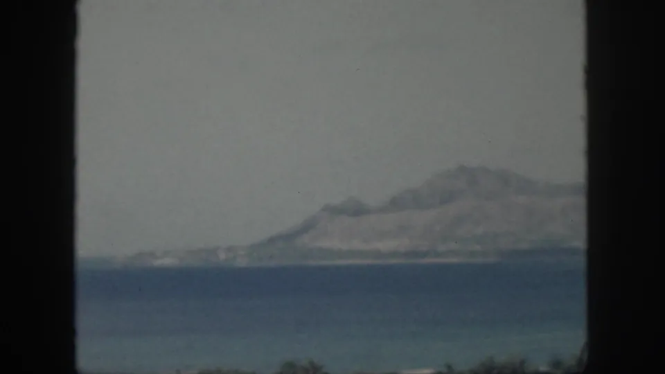 1959 Oahu home-movie frame showing a distant mountainous coast across the water, bordered by a dark vertical window or frame edge.