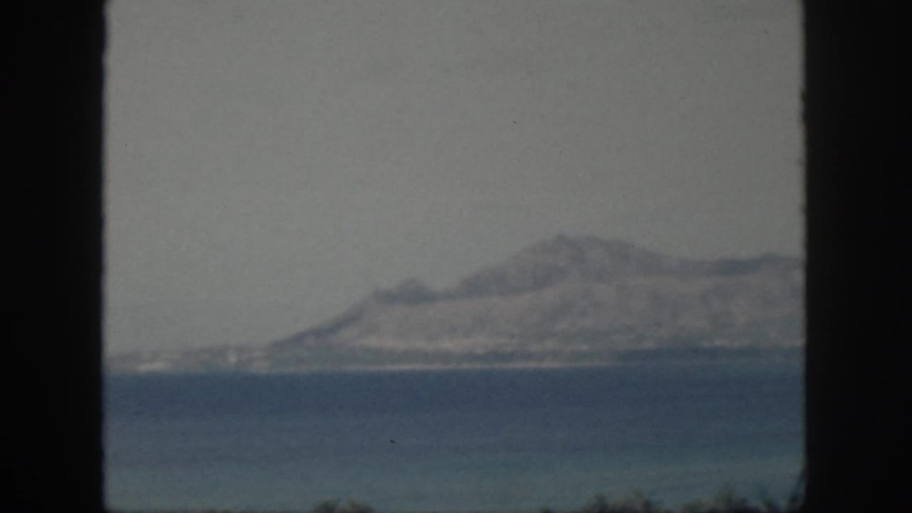 1959 Oahu home-movie frame showing a hazy mountainous island across the sea through a dark window opening.