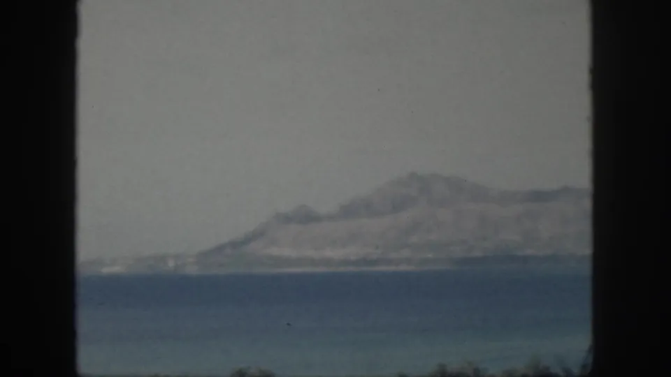 1959 Oahu home-movie frame showing a hazy mountainous island across the sea through a dark window opening.