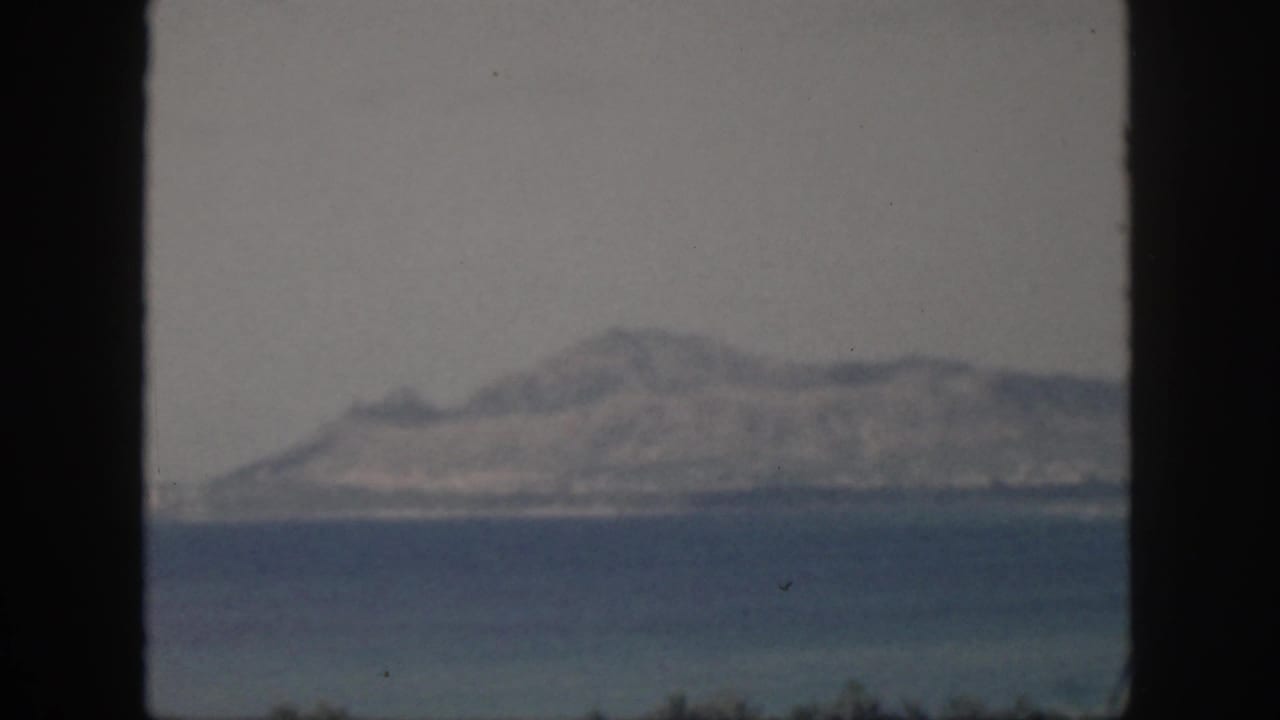 Soft 8mm view from Oahu in 1959, with a distant island ridge, hazy sky, and blue water divided by a window frame.