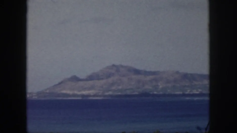 1959 Hawaii travel footage frame with a rugged island silhouette across blue water, partly bordered by a dark window edge.