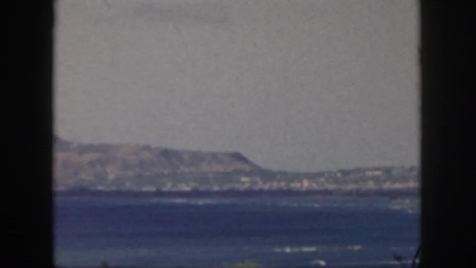 Vintage 8mm Oahu coastal view with surf streaks, distant shore, and muted hillside under haze.