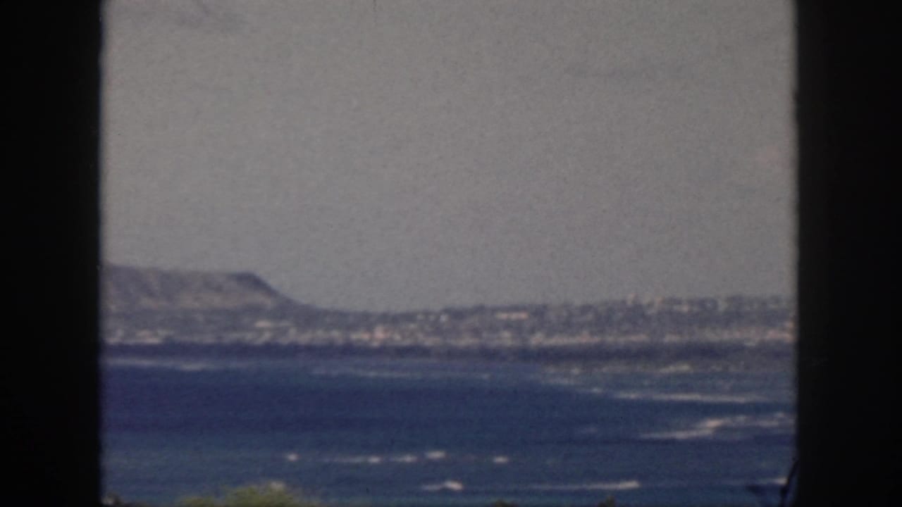 1959 Oahu coastal view with blue ocean, white surf, hazy hills, and a dark window frame dividing the scene.